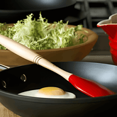 Espátula pequena de silicone vermelha Le Creuset em uso com frigideira e ovo frito. Ao fundo, bowl com folhas verdes. Ideal para quem busca praticidade na cozinha gourmet, combinando com porta temperos, bandeja e itens de mesa posta.